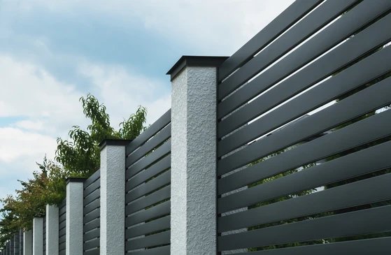 Scène extérieure avec clôture moderne à lames horizontales perforées en métal gris, laissant apercevoir la végétation au travers